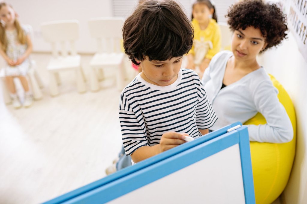 a-pontoise-l-inclusion-scolaire-face-au-manque-de-moyens Un garçon écrit sur un tableau blanc encadré de bleu, sous le regard d’une femme assise, avec d’autres enfants en