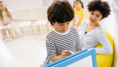 Un garçon écrit sur un tableau blanc encadré de bleu, sous le regard d’une femme assise, avec d’autres enfants en